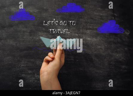 Hand holding a paper boat on a chalkboard with the words Let's go to travel written below it Stock Photo