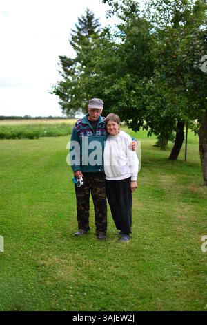 Senior couple standing together in rural area in summer day happily smiling. Elderly man hugs his elderly wife in countryside. Casual village scene po Stock Photo