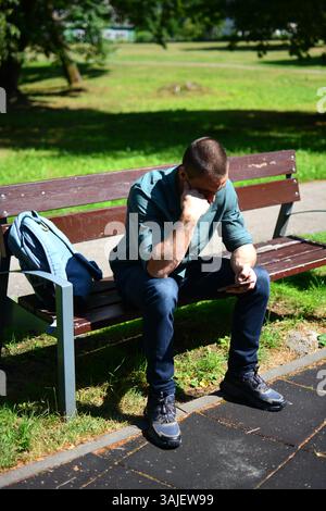 Man in green shirts is scrolling phone and sitting on a bench outside in hot sunny day Stock Photo