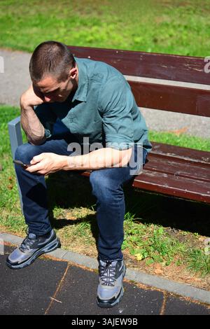 Man in green shirts is scrolling phone and sitting on a bench outside in hot sunny day Stock Photo