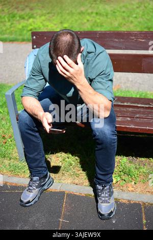 Man in green shirts is scrolling phone and sitting on a bench outside in hot sunny day Stock Photo