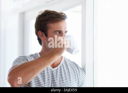 Man, window and drinking coffee for relaxing at home, hot tea and beverage for contemplating. Morning routine, male person and daydreaming in Stock Photo