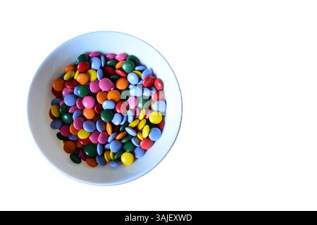 A full bowl of colourful candies top view isolated background. Chocolate buttons sweet treats in white bowl. Stock Photo