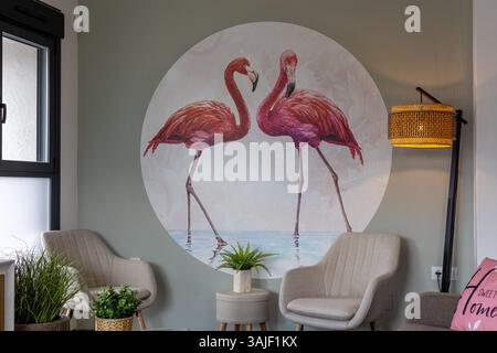 A bright and stylish seating area featuring two beige chairs, a stool with a plant, and a bold flamingo mural on the wall, creating a cozy and tropica Stock Photo