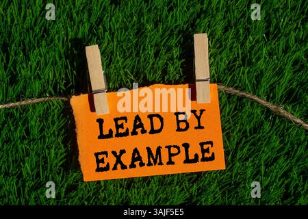 Lead by example message written on ripped torn orange paper piece on green grass background. Conceptual lead by example symbol. Copy space. Stock Photo