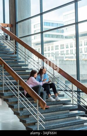 Two students studying using a laptop while sitting on the stairs in the university lobby Stock Photo