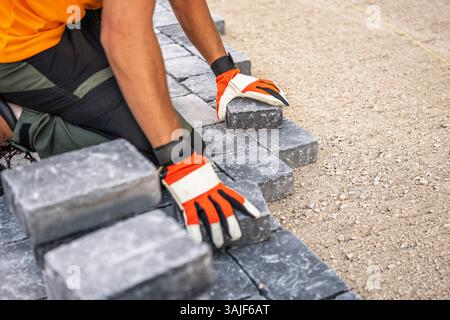 A construction worker is carefully laying gray stone blocks on a prepared surface. The worker, wearing an orange shirt and gloves, focuses on aligning Stock Photo