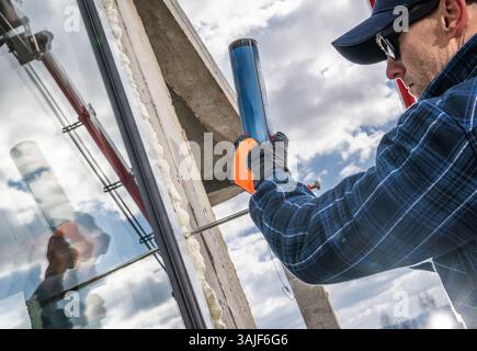 A construction worker applies sealant to install glass in a building while using a caulking gun. The cloudy sky serves as a backdrop, highlighting the Stock Photo