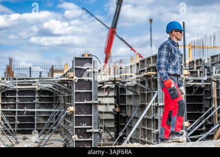 A construction worker in a helmet and checkered shirt surveys a construction site filled with scaffolding and forming systems. Cranes are visible in t Stock Photo