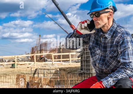 A construction worker in a blue helmet and protective gear is sitting on a site while talking on his smartphone. Behind him, cranes and construction m Stock Photo