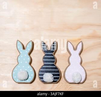 Three small rabbits with white pom poms on their tails are sitting on a wooden surface. The rabbits are of different colors, with one being blue, one Stock Photo