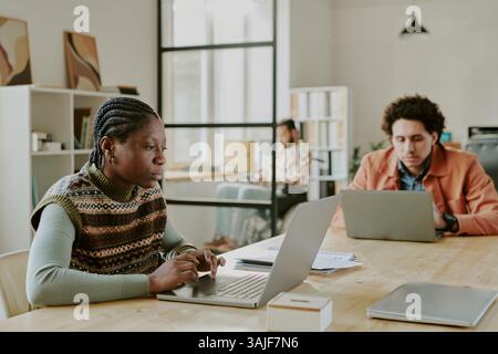 Medium shot of serious and concentrated African American woman with tiny braids looking at laptops screen while programming on it, her curly haired male colleagues coding near her Stock Photo