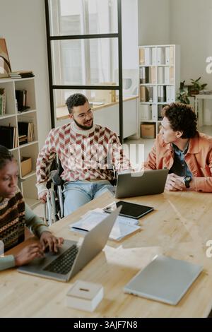 Vertical high angle shot of three biracial young adult programmers working at wooden table in center of spacious office, two men discussing new app together Stock Photo