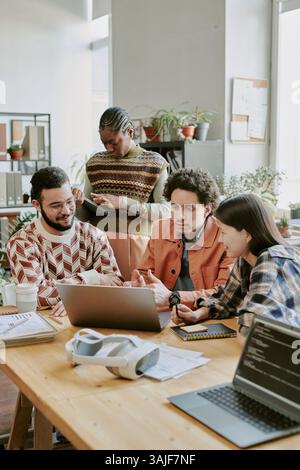 Vertical shot of biracial team of app developers sitting at table and looking at screen of laptop while having informal meeting Stock Photo