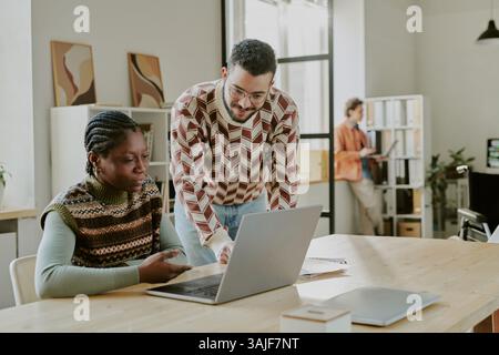 Medium shot of handsome young male senior developer helping his African American female junior colleague with codes on laptop Stock Photo