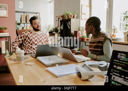 Biracial male and female programmers chatting while sitting at wooden desk with laptops, copybooks and documents on top Stock Photo