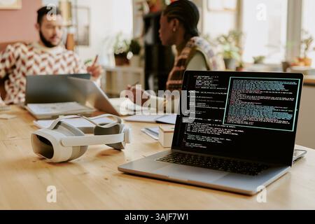 Medium close up of opened silver laptop with code lines on screen and headphones on top of desk, biracial colleagues interaction with each other in blurred background Stock Photo
