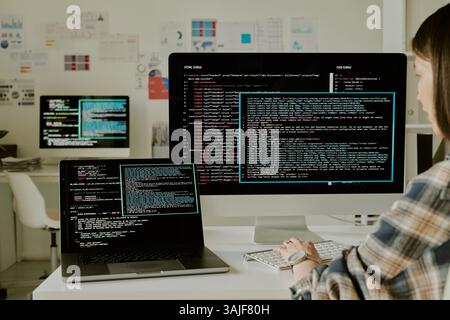 Medium shot of unrecognizable female programmer coding on computer in office, her workplace filled with digital devices with huge monitors Stock Photo