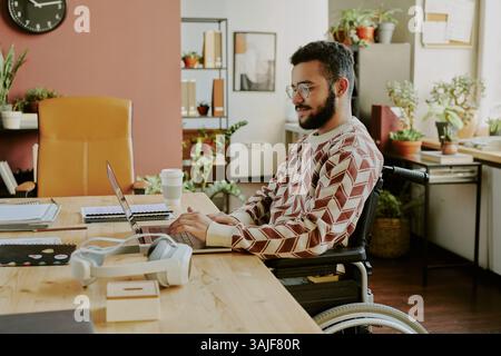 Medium shot of highly skilled programmer with disability working on laptop in office decorated with huge number of fresh green plants Stock Photo