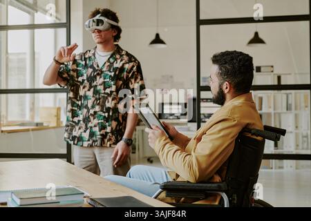 Medium shot of male talented and highly skilled gamedevs testing new project together in office, curly haired man wearing VR set while his coworker controlling game process on touchpad Stock Photo