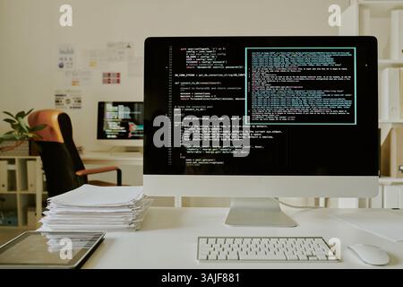 Medium close up of computer with code lines on monitor, stack of documents and tablet on top of desk in office Stock Photo