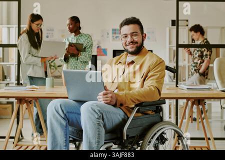 Portrait of handsome young IT specialist with disability holding laptop of knees smiling at camera while his colleagues working in background Stock Photo