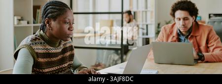 Website banner with biracial young adult coworkers sitting at desk, looking at laptops monitor during workday in comfortable office Stock Photo