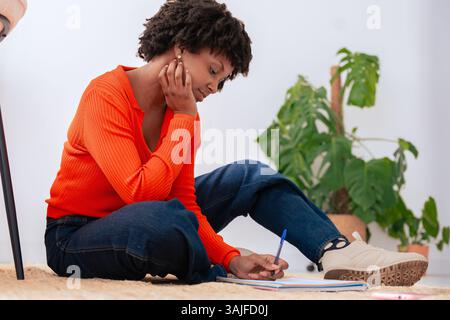 African american woman sitting on floor writing in notebook cozy home interior Stock Photo