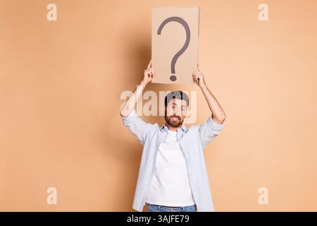 Young Man Holding a Question Mark Poster Against Beige Background in Casual Attire Stock Photo