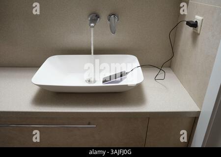 Smartphone connected to power outlet submerged in water inside bathroom sink with running tap. Studio shot of electrical hazard and water damage risk. Stock Photo