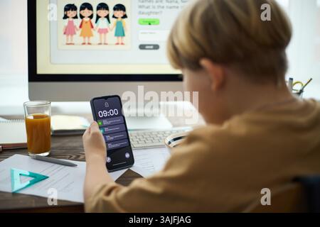 Boy checking time on smartphone while doing school homework, with computer screen, notepad, and glass of orange juice on desk Stock Photo