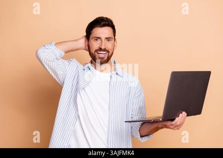 Young man with laptop smiling confidently in beige background wearing casual striped shirt Stock Photo