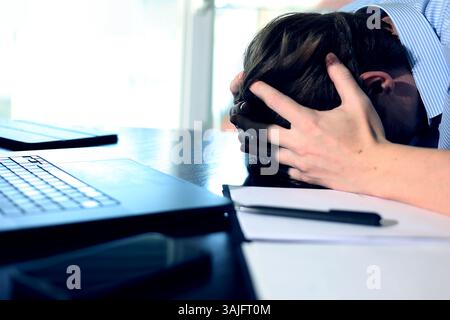 man at computer holding his head, work online. a man is working on a laptop, there is a notebook and a pen lying next to him, he grabbed his head Stock Photo