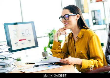 Skilled businesswoman working on project at modern office desk while analyzing charts and data, a professional workspace setting Stock Photo