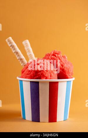 Strawberry ice cream in striped paper cup with wafer sticks. Vibrant summer dessert in red and orange tones on minimal studio background. Stock Photo