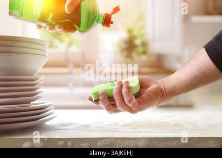 Person washing dishes in kitchen pouring dishwashing product from a bottle with green detergent onto a scouring pad on a white counter with a stack of Stock Photo
