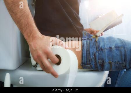 A man sitting on a toilet bowl with, defecating with a roll of white toilet paper in one hand and with a book reading on the other. Stock Photo
