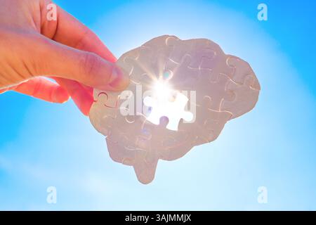 Hand holds a white puzzle piece shaped like a human brain against a bright blue sky, with sunlight shining through the missing piece opening. Mental h Stock Photo