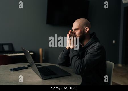 Side view of exhausted freelancer male covering face with hand while sitting at desk in home office, feeling tired and overwhelmed by work. Stock Photo