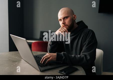 Portrait of focused freelancer sitting at table, working on laptop computer while resting hand on chin, deep in thought looking at camera Stock Photo
