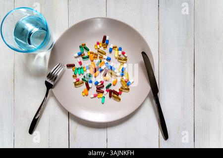 Mixed pills on plate with fork and knife. Various colorful capsules and tablets are scattered on a plate, styled like a meal, with cutlery and a glass Stock Photo