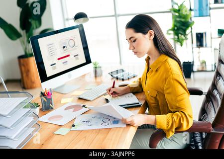 Skilled professional woman analyzing data at a modern office workspace with computer and documents during a bright daylight setting Stock Photo
