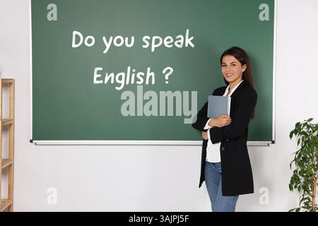 Teacher near green chalkboard with phrase Do You Speak English? in classroom Stock Photo