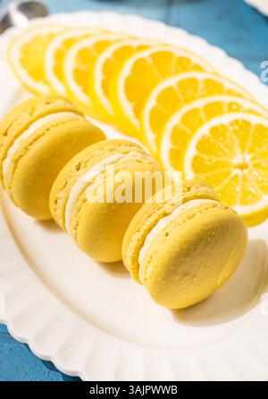 Delicious lemon-flavored macarons arranged artfully on a white plate, complemented by fresh lemon slices, set against a vibrant blue table Stock Photo