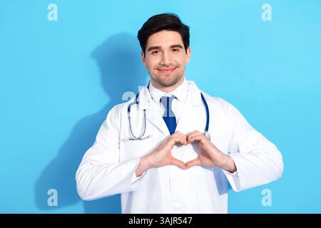 Smiling young doctor in white coat forming heart shape with hands against blue background showcasing compassion for healthcare Stock Photo