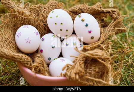 A close-up of white Easter eggs adorned with colorful sequins and arranged in a rustic jute basket on green grass, perfect for spring or Easter-themed Stock Photo