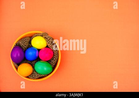 Colorful Easter Eggs in Bowl with Rustic Thread Decor isolated On orange. Multiple bright Easter eggs placed inside an orange bowl filled with wooden Stock Photo