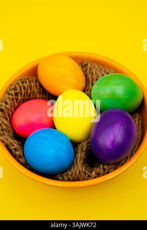 Vibrant painted Easter eggs placed inside an orange bowl filled with rustic wooden thread over a cheerful yellow surface. Stock Photo