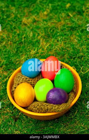 Vibrant painted Easter eggs placed inside an orange bowl filled with rustic wooden thread on grass. Stock Photo