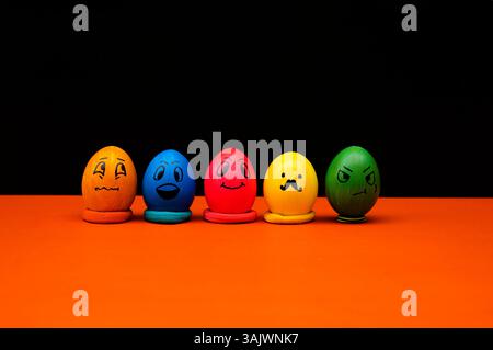 Five brightly colored eggs with distinct hand-drawn expressions, representing various emotions. Set against an orange surface and black background, th Stock Photo